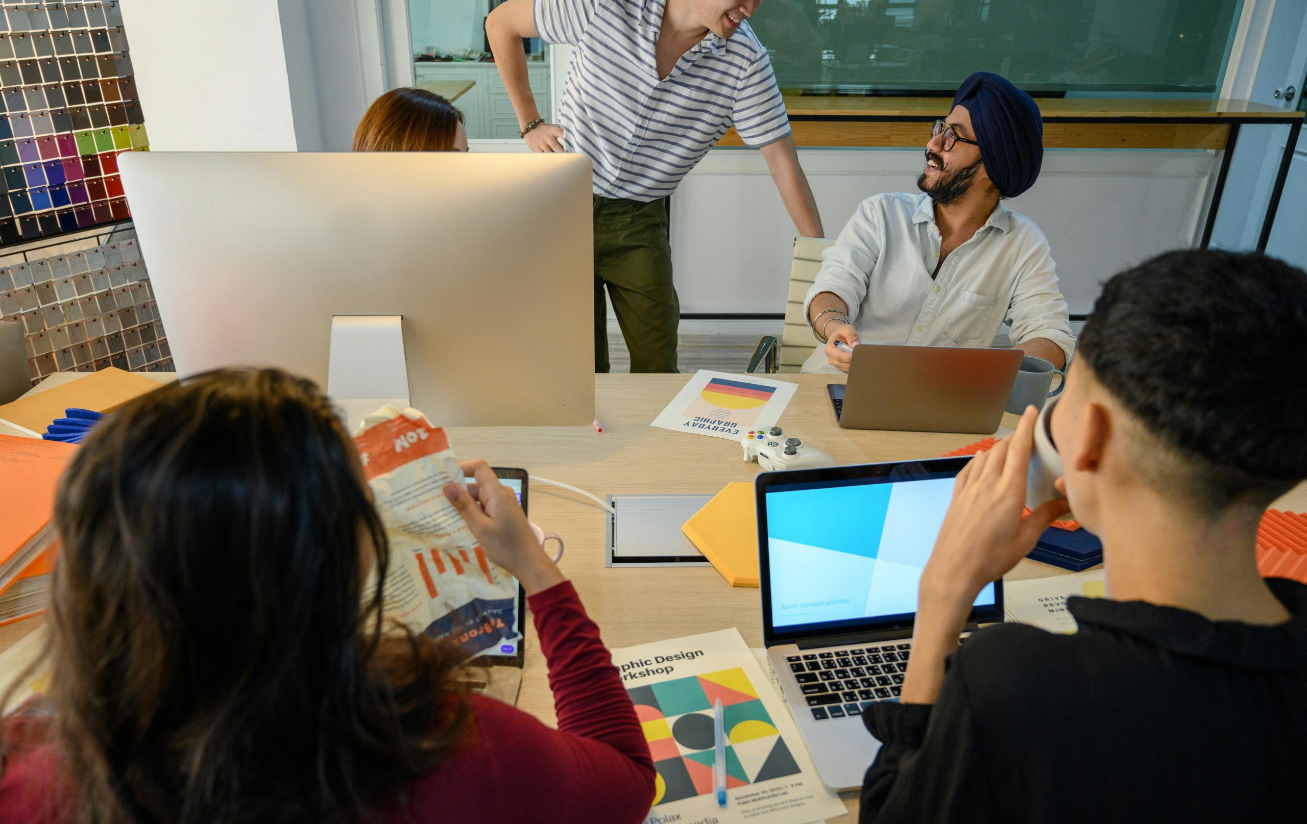 A small creative team collaborates around a table in a modern design studio, reviewing laptops, printed workshop materials and colour samples during a branding or graphic design discussion.