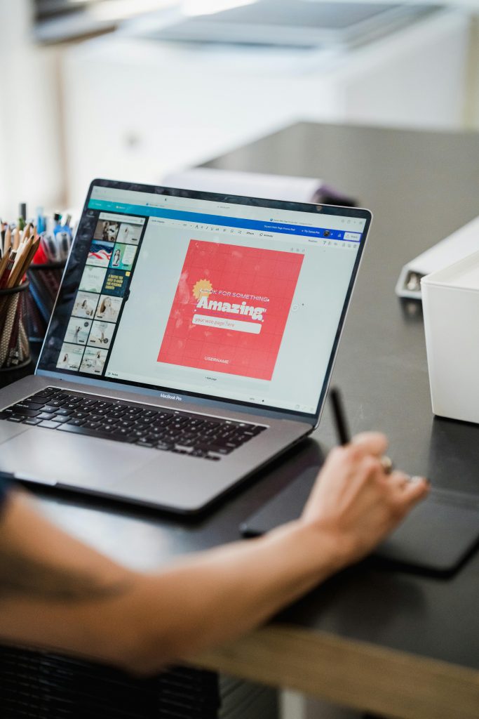 A designer uses a drawing tablet beside a laptop displaying a red poster design that reads “Look for Something Amazing,” surrounded by template thumbnails and coloured pencils on the desk.